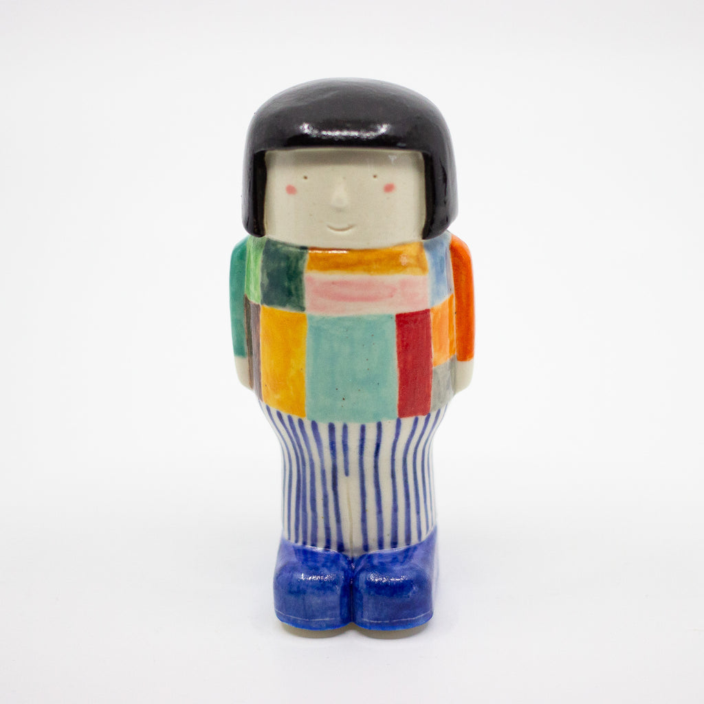 Front view of a handmade ceramic figurine with a black bob haircut, smiling face, colourful patchwork top, vertical striped pants, and blue shoes. Designed by shuh.