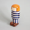 Ceramic figurine of a person with orange hair, striped blue and white outfit, and brown shoes on a gray background