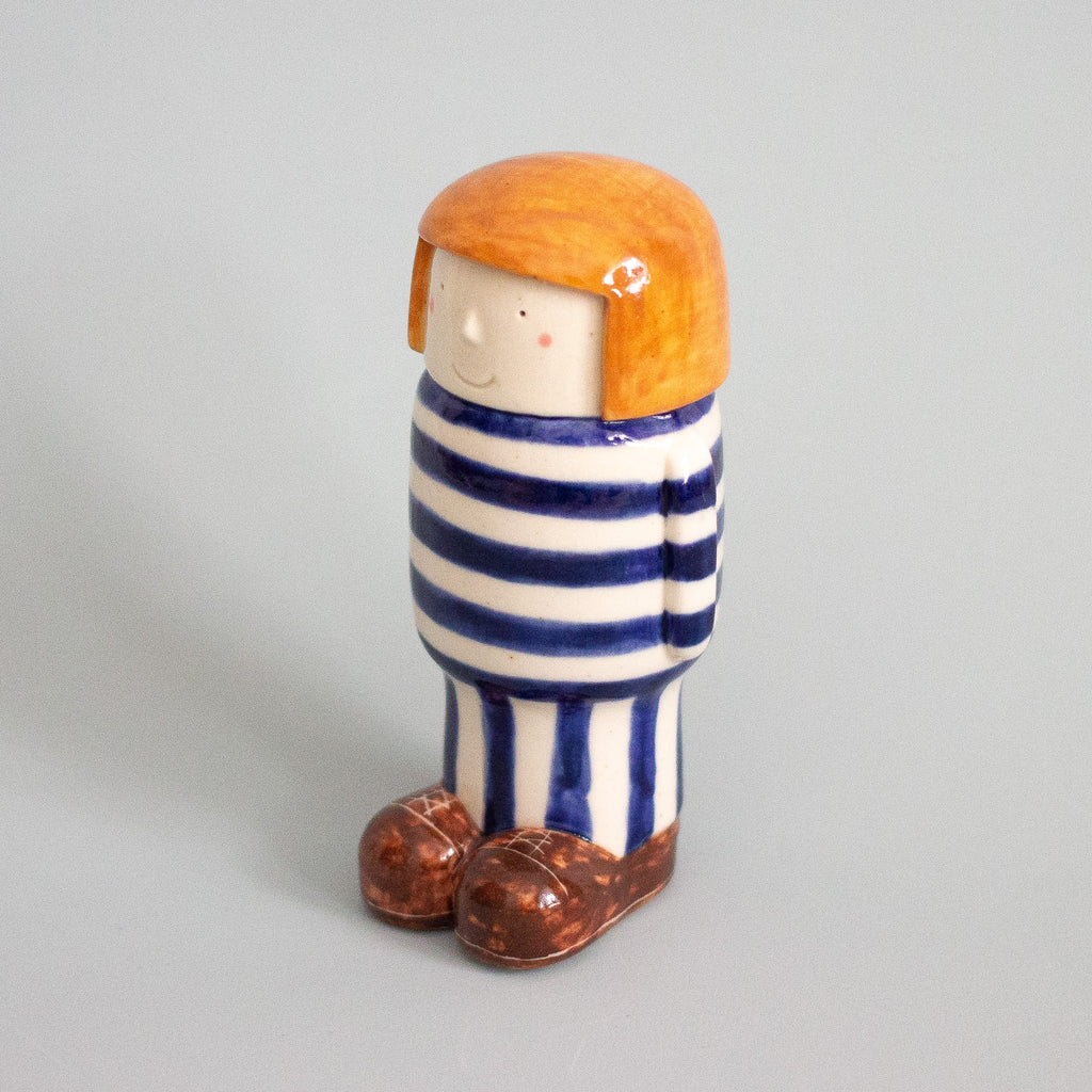 Ceramic figurine of a person with orange hair, striped blue and white outfit, and brown shoes on a gray background