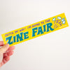 A hand holding a bright yellow rectangular sticker that reads 'OUTTA MY WAY! I'M GOING TO THE ZINE FAIR' in teal and purple lettering, with small white line drawings and 'Helio Press' printed on the right side, against a plain light background.