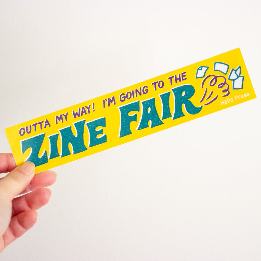 A hand holding a bright yellow rectangular sticker that reads 'OUTTA MY WAY! I'M GOING TO THE ZINE FAIR' in teal and purple lettering, with small white line drawings and 'Helio Press' printed on the right side, against a plain light background.