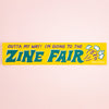 A rectangular yellow bumper sticker with the text 'OUTTA MY WAY! I'M GOING TO THE ZINE FAIR' in bold teal and purple lettering, plus small illustrated magazine covers and the words 'Helio Press' on the right, shown against a pale pink background.