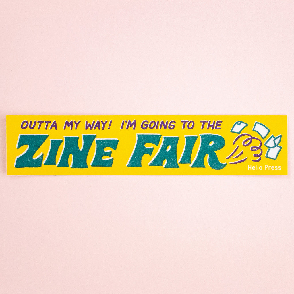 A rectangular yellow bumper sticker with the text 'OUTTA MY WAY! I'M GOING TO THE ZINE FAIR' in bold teal and purple lettering, plus small illustrated magazine covers and the words 'Helio Press' on the right, shown against a pale pink background.