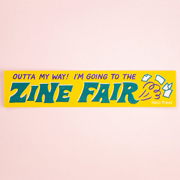 A rectangular yellow bumper sticker with the text 'OUTTA MY WAY! I'M GOING TO THE ZINE FAIR' in bold teal and purple lettering, plus small illustrated magazine covers and the words 'Helio Press' on the right, shown against a pale pink background.