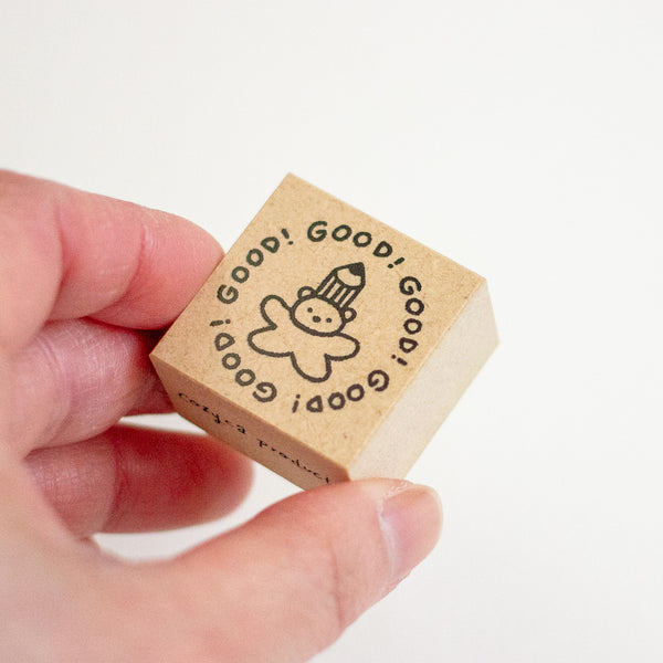 A hand holding a small square wooden stamp block with black printed text reading 'Good! Good! Good!' and a simple cartoon face, against a plain white background.