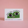 A white greeting card featuring a green cartoon face with large black eyes and the text 'HELLO, I QUITE LIKE YOUR FACE', photographed against a soft pink background.