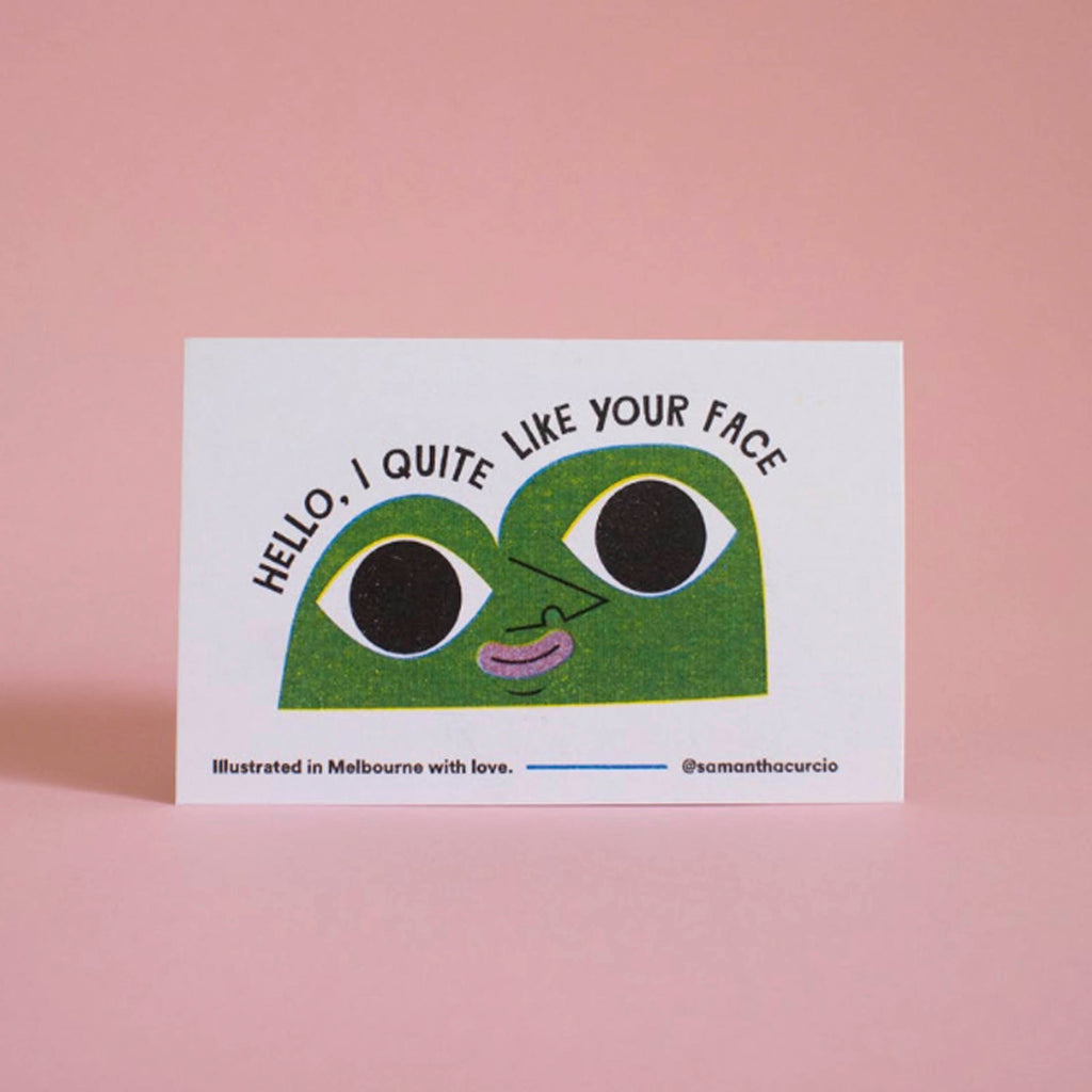 A white greeting card featuring a green cartoon face with large black eyes and the text 'HELLO, I QUITE LIKE YOUR FACE', photographed against a soft pink background.