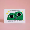 A green embroidered iron-on patch shaped like a smiling heart with black eyes and a purple mouth, displayed on a white backing card that reads 'Samantha Curcio.' and 'GREEN BOI IRON-ON PATCH,' against a soft pink background.