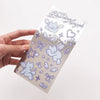 A hand holds a clear metallic silver sticker sheet decorated with cute purple angel illustrations, including winged cats, bows, stars, and hearts, against a plain white background.