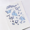 A sheet of cute die-cut stickers featuring light blue cherubs, hearts, stars, and bows with the text 'Guardian Angels' in blue script, displayed on an open white notebook.