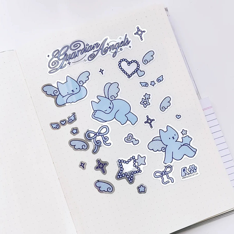 A sheet of cute die-cut stickers featuring light blue cherubs, hearts, stars, and bows with the text 'Guardian Angels' in blue script, displayed on an open white notebook.
