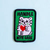 An embroidered rectangular patch with a green background, black stitched border, black text reading 'HANDLE WITH CARE,' and a white cartoon holding a red heart.