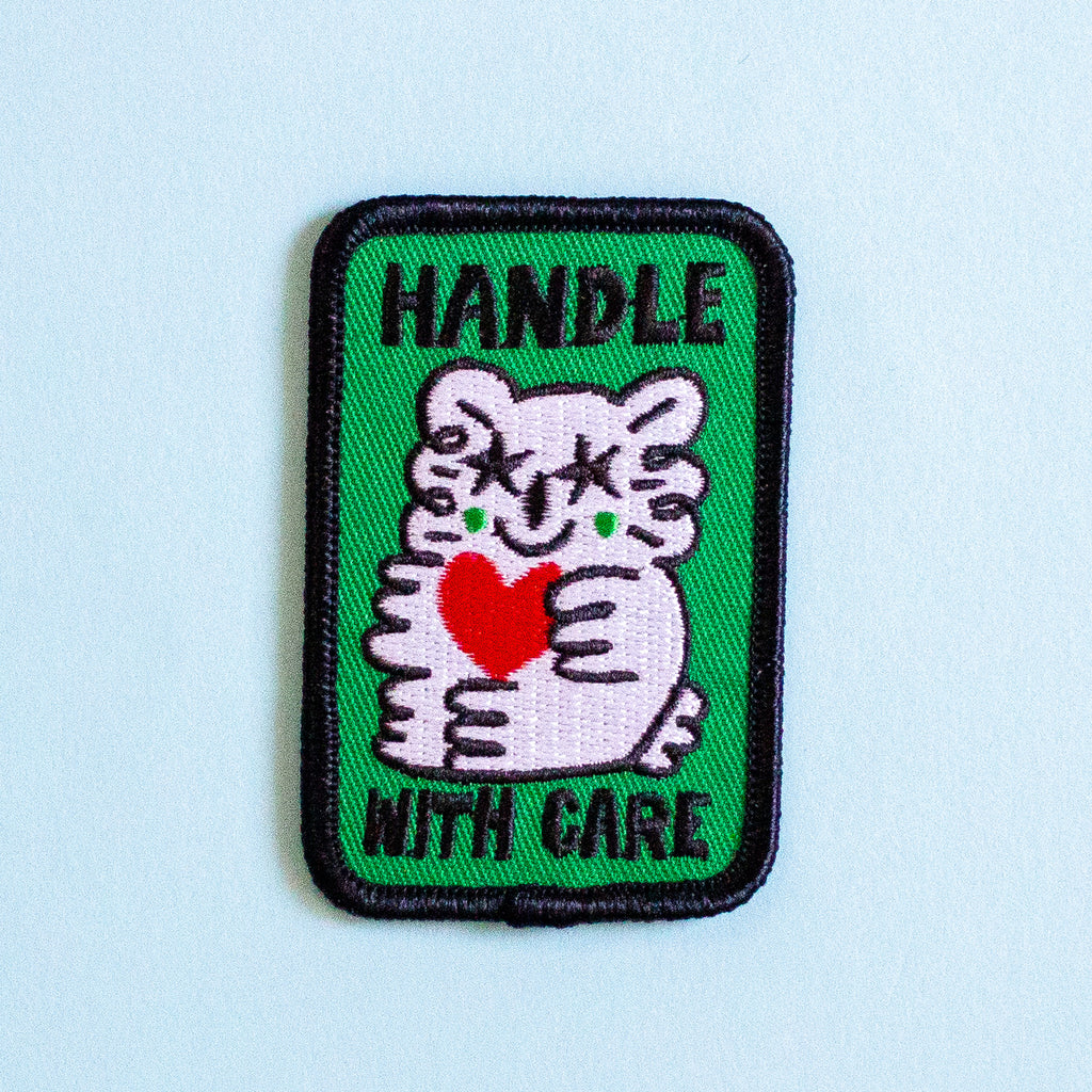 An embroidered rectangular patch with a green background, black stitched border, black text reading 'HANDLE WITH CARE,' and a white cartoon holding a red heart.