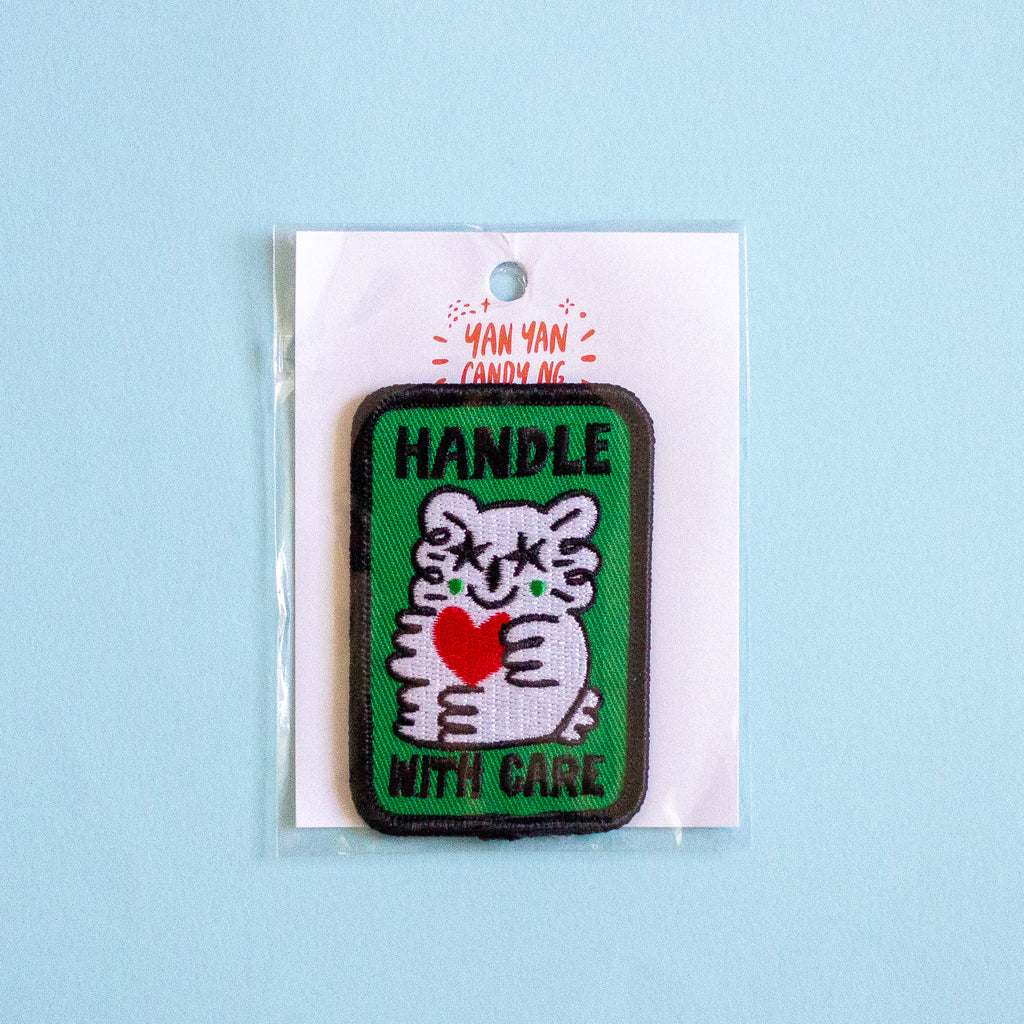 An embroidered patch featuring a white cartoon holding a red heart on a green background with the text 'HANDLE WITH CARE' in black. The patch is displayed on a white backing card with the brand name 'YAN YAN CANDY NG' printed in red, set against a light blue background.