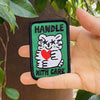 A hand holds a rectangular embroidered patch featuring a white cartoon hugging a red heart, with the text 'HANDLE WITH CARE' on a green background, set against a background of green leaves and a brown tree trunk.