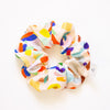 Colourful fabric scrunchie with a painterly abstract print in red, blue, yellow, green, and pastel tones on a light background, displayed in a circular shape on a white surface.