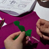 A close-up of hands crafting with felt shapes and scissors on a white table, with an instruction sheet visible in the background.