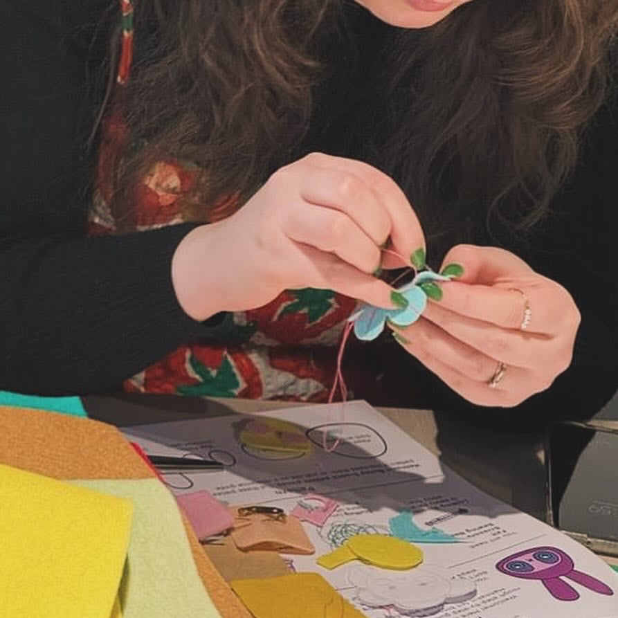 A person crafting with colourful felt pieces on a table, holding a small blue and green felt flower. Papers with illustrations, scissors, a phone, and felt sheets are visible on the tabletop.