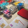 A tabletop display of colourful felt craft supplies, including clear plastic tubs filled with turquoise, pink, yellow, blue, black, and white felt pieces, a large red felt sheet, and printed activity sheets with bunny illustrations and small cards on a light wood table.