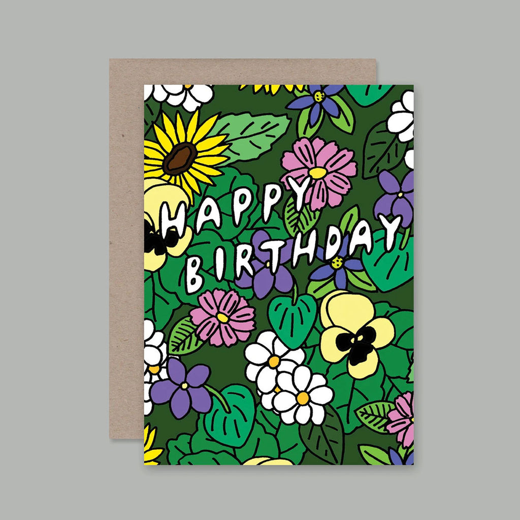 A greeting card with a colourful floral pattern and the words “HAPPY BIRTHDAY” in white outlined lettering, shown with a plain kraft envelope behind it on a neutral grey background.