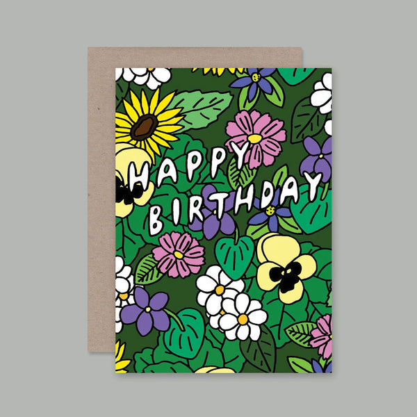 A greeting card with a colourful floral pattern and the words “HAPPY BIRTHDAY” in white outlined lettering, shown with a plain kraft envelope behind it on a neutral grey background.