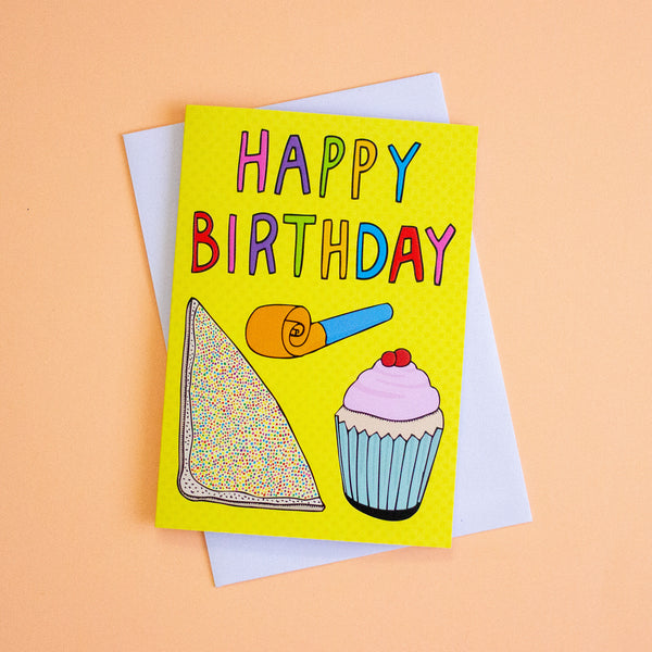 A bright yellow birthday card with colourful 'HAPPY BIRTHDAY' lettering and illustrations of a slice of cake, a party popper, and a cupcake, shown with a white envelope on a peach background.