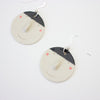 An angled view of a pair of handmade ceramic earrings by shuh. featuring smiling faces with raised noses, rosy cheeks, and black hair, finished with silver hooks and displayed on a white background.