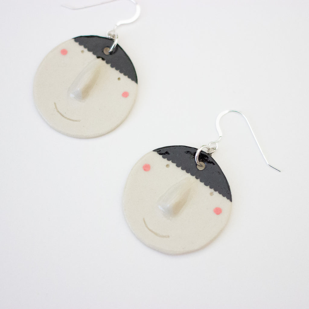 An angled view of a pair of handmade ceramic earrings by shuh. featuring smiling faces with raised noses, rosy cheeks, and black hair, finished with silver hooks and displayed on a white background.