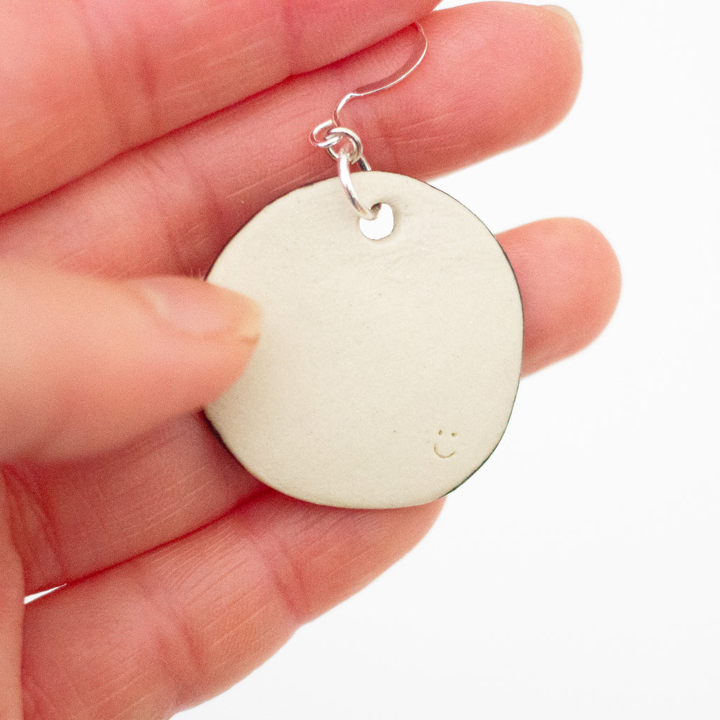 Photo of the back of the happy face handmade ceramic earrings, showing plain ceramic backing with small carved smiley face. 