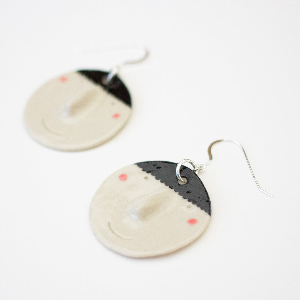 Side view of a pair of handmade ceramic earrings by shuh. featuring smiling faces with raised noses, rosy cheeks, and black hair, finished with silver hooks and displayed on a white background.