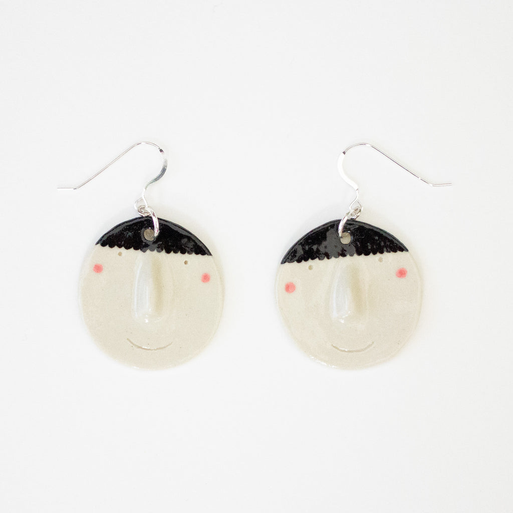 Pair of handmade ceramic earrings by shuh. featuring smiling faces with raised noses, rosy cheeks, and black hair, finished with silver hooks and displayed on a white background.
