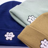 Close-up of three knit beanies in navy blue, sage green, and mustard yellow, each featuring an embroidered smiling flower patch on the front fold, showing texture and detail of the fabric and stitching.