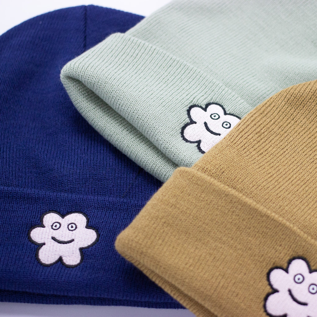 Close-up of three knit beanies in navy blue, sage green, and mustard yellow, each featuring an embroidered smiling flower patch on the front fold, showing texture and detail of the fabric and stitching.
