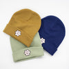 Three knit beanies in mustard yellow, sage green, and navy blue, each featuring an embroidered smiling flower patch on the front fold, arranged in a fan-like layout on a white background.