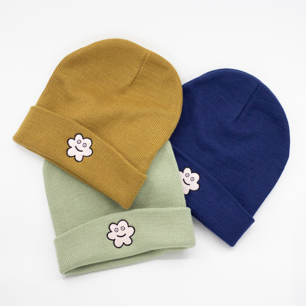 Three knit beanies in mustard yellow, sage green, and navy blue, each featuring an embroidered smiling flower patch on the front fold, arranged in a fan-like layout on a white background.