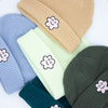 A group of wide knit beanies in soft textured fabric, shown in tan, pine green, and lime green, each with a folded brim and a smiling embroidered flower patch on the front, arranged in a colourful flat lay.