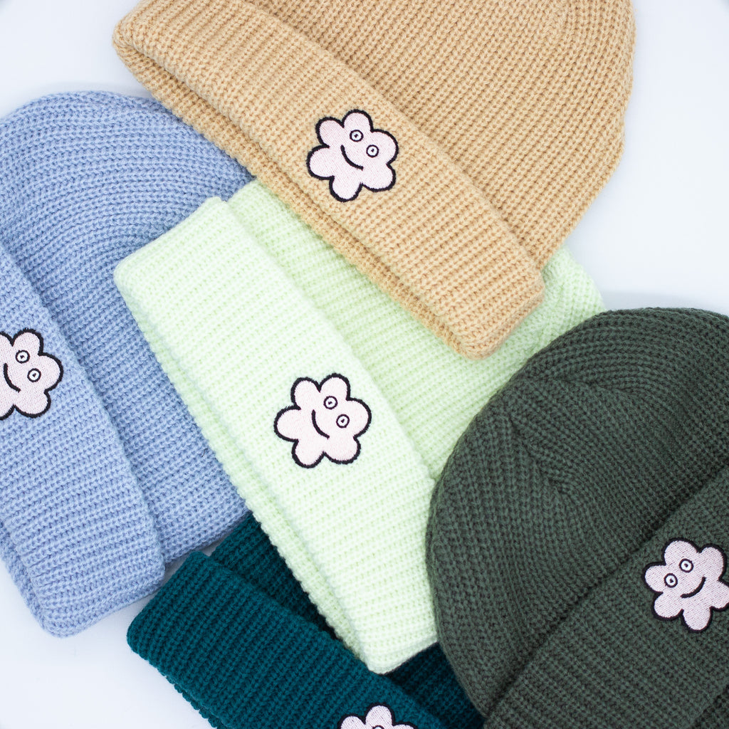 A group of wide knit beanies in soft textured fabric, shown in tan, pine green, and lime green, each with a folded brim and a smiling embroidered flower patch on the front, arranged in a colourful flat lay.
