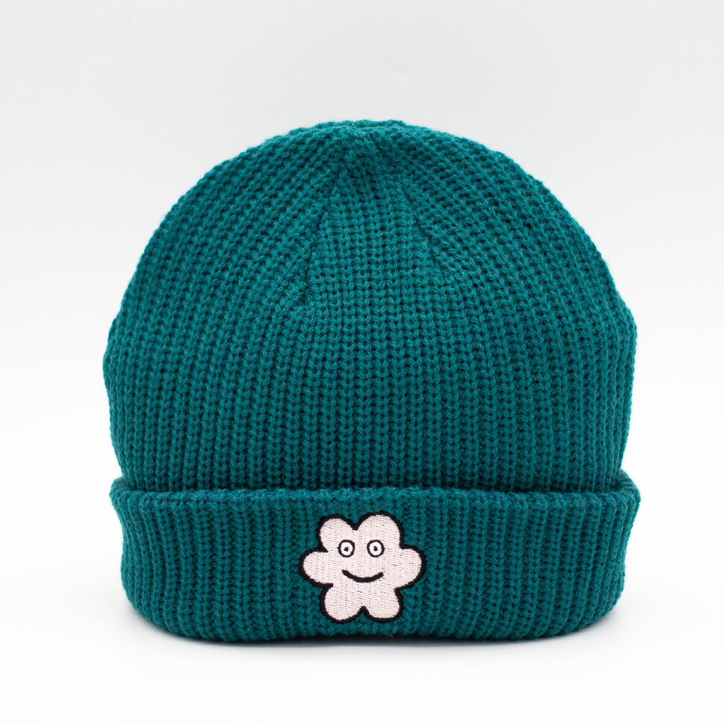 Front view of a Pine wide knit beanie with a folded brim, featuring a smiling embroidered flower patch on the front, displayed against a plain white background.