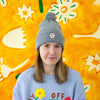 Person wearing a gray beanie with a flower design and a delightful pom pom. Also wearing a light blue sweatshirt with colorful graphics against a bright yellow background with abstract shapes.