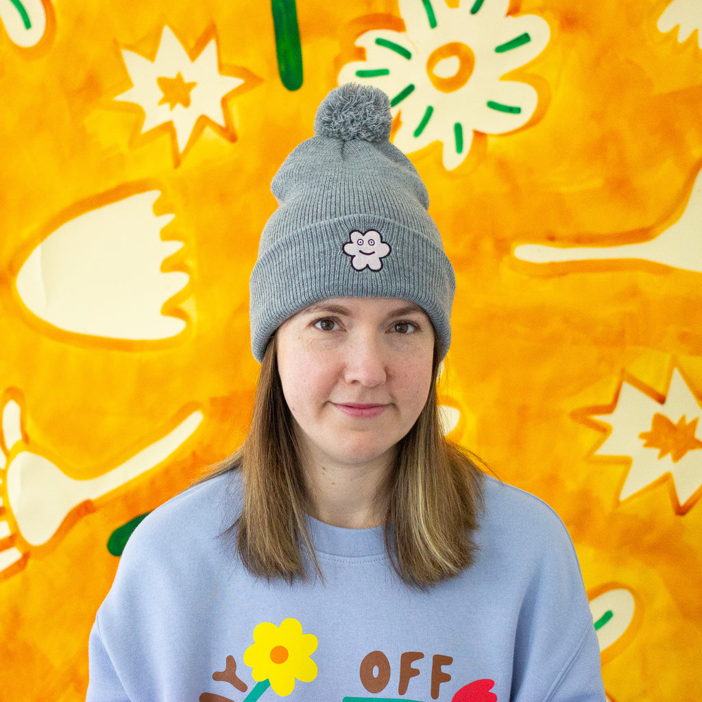 Person wearing a gray beanie with a flower design and a delightful pom pom. Also wearing a light blue sweatshirt with colorful graphics against a bright yellow background with abstract shapes.
