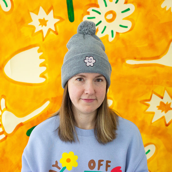 Person wearing a gray beanie with a flower design and a delightful pom pom. Also wearing a light blue sweatshirt with colorful graphics against a bright yellow background with abstract shapes.