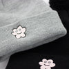 Close-up of a grey and a black knit beanie, each featuring a stitched smiling flower patch on the front fold, with visible knit texture and a partial view of the pom-poms at the top.