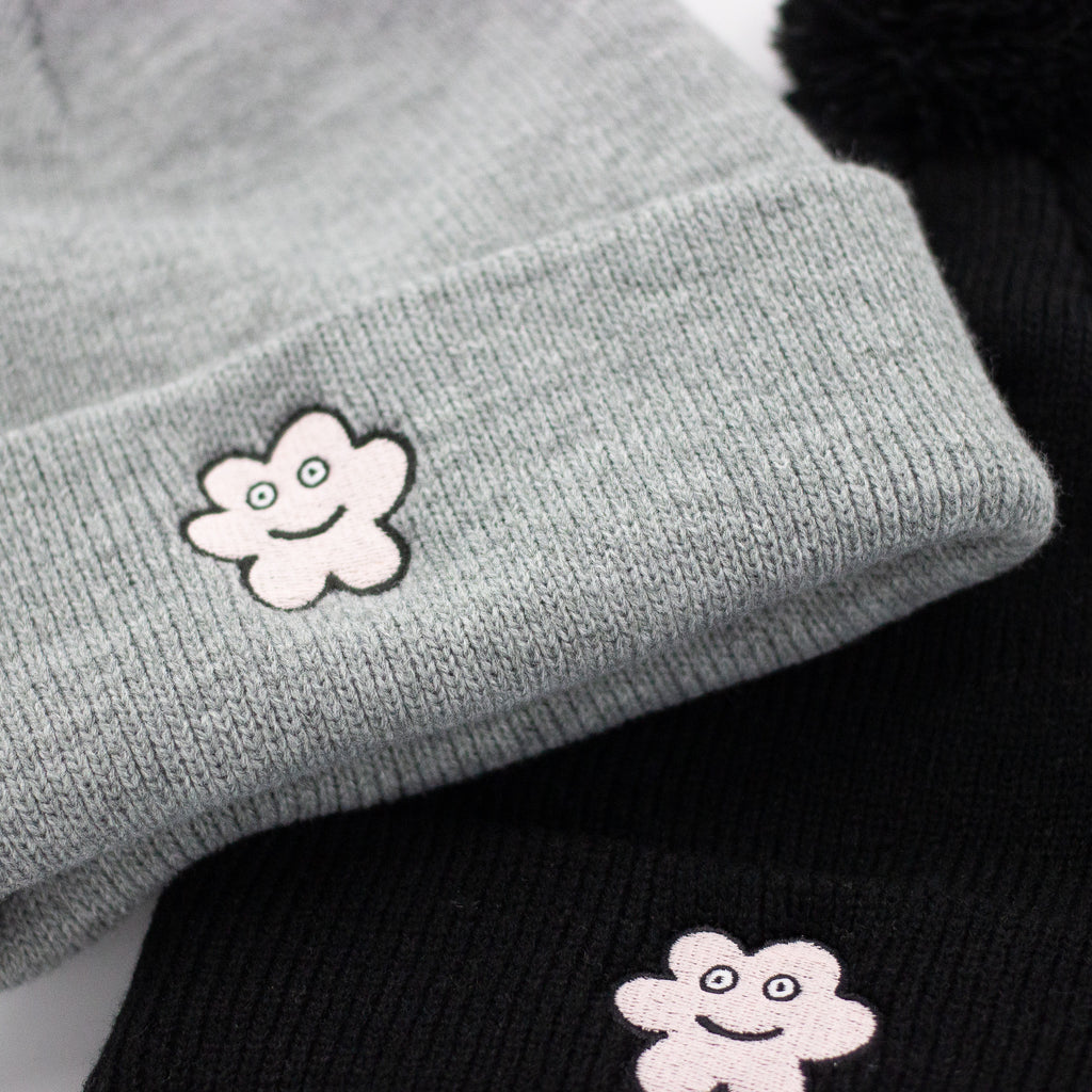 Close-up of a grey and a black knit beanie, each featuring a stitched smiling flower patch on the front fold, with visible knit texture and a partial view of the pom-poms at the top.