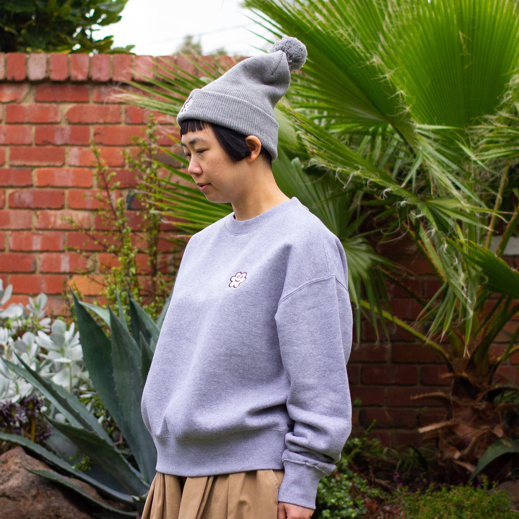 Person standing outdoors in front of a brick wall and lush green plants, wearing a grey pom-pom beanie and a matching grey sweatshirt, both featuring a small embroidered smiling flower patch.