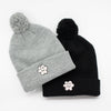 Two knit beanies, one in grey and one in black, each with a fluffy pom-pom on top and an embroidered smiling flower patch on the front fold, displayed side by side on a white background.