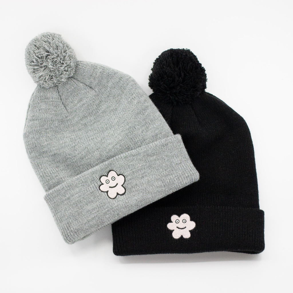 Two knit beanies, one in grey and one in black, each with a fluffy pom-pom on top and an embroidered smiling flower patch on the front fold, displayed side by side on a white background.