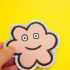 A hand holding a die-cut sticker featuring a cute flower shaped character with a holographic border, set against a plain yellow background.