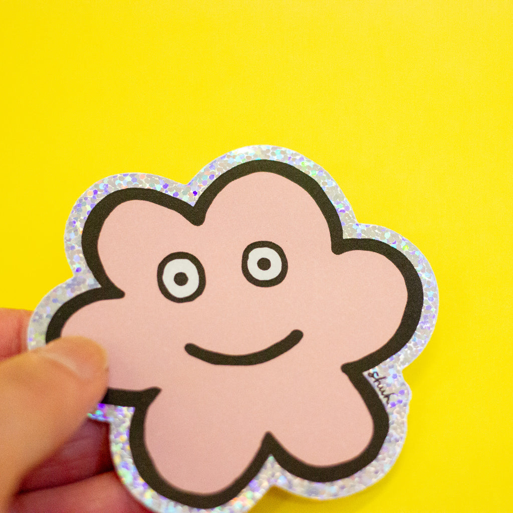 A hand holding a die-cut sticker featuring a cute flower shaped character with a holographic border, set against a plain yellow background.