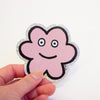 A hand holding a die-cut sticker featuring a cute flower shaped character with a holographic border, set against a plain white background.