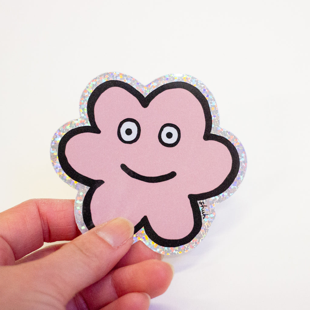 A hand holding a die-cut sticker featuring a cute flower shaped character with a holographic border, set against a plain white background.